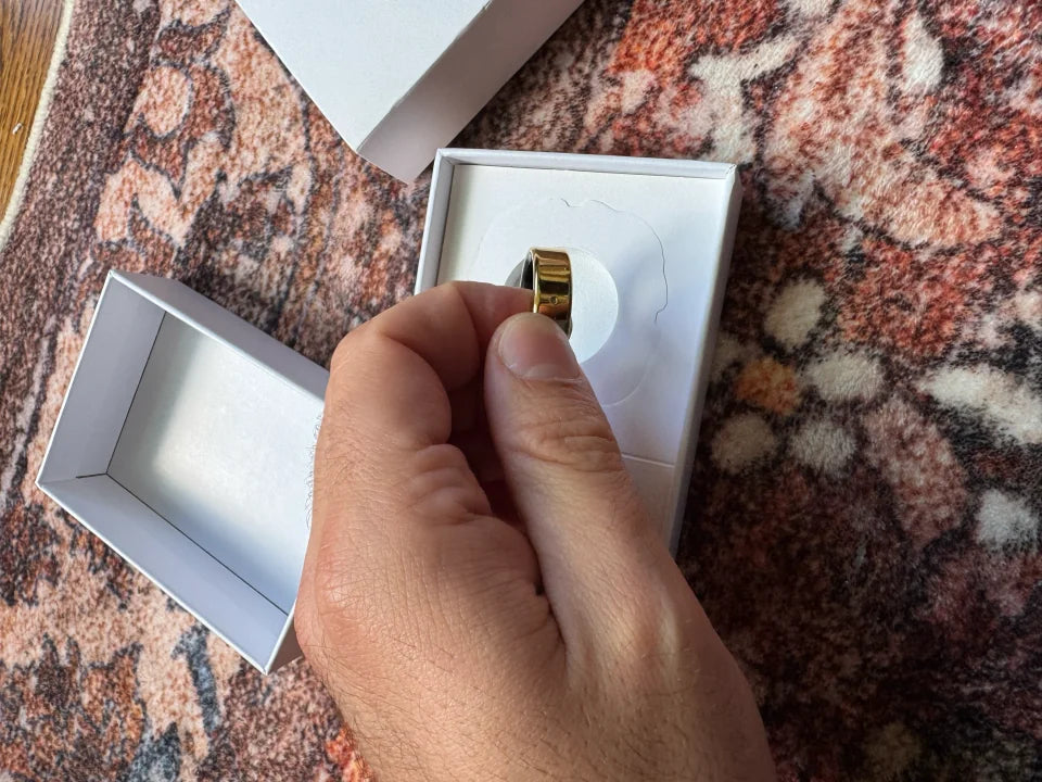 A hand lifts a gold Secondly ring from its white box on a patterned rug.