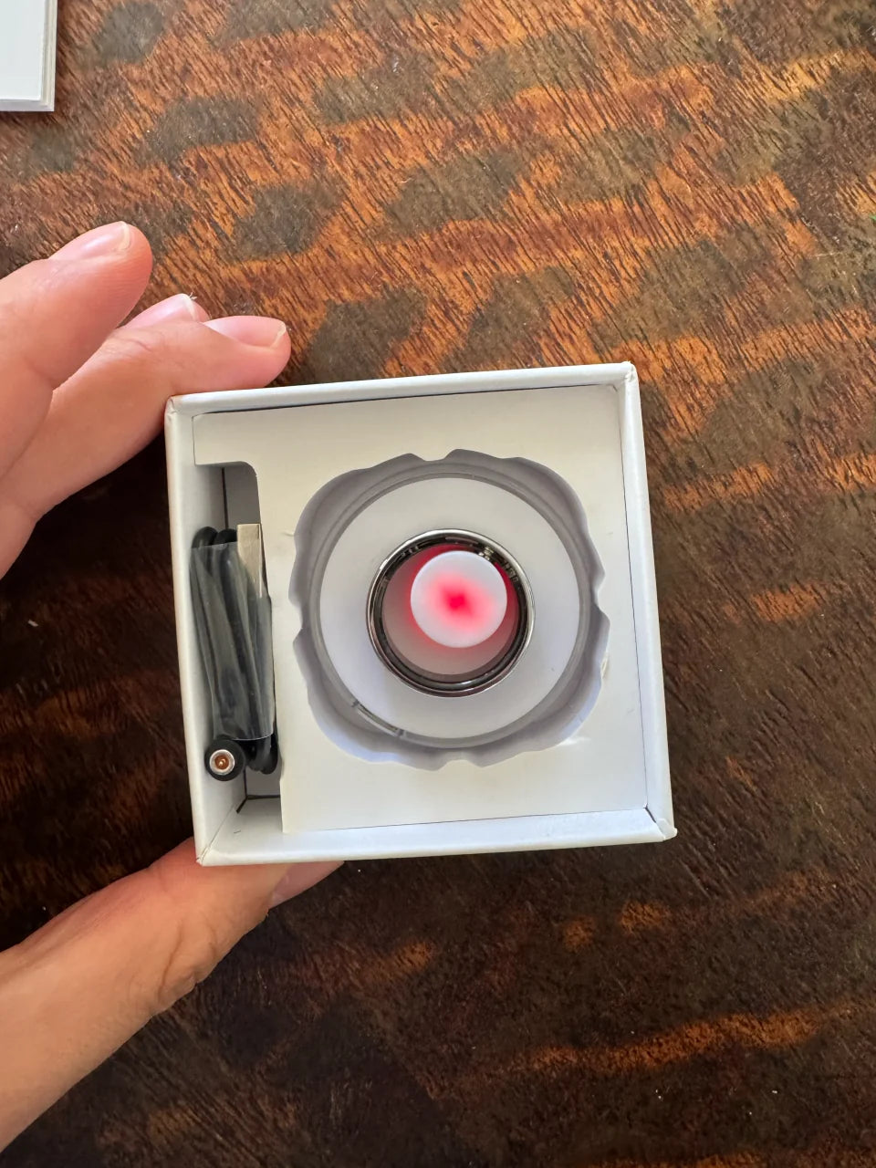 An overhead view of a silver Secondly ring and cable inside an open box on a wooden table.
