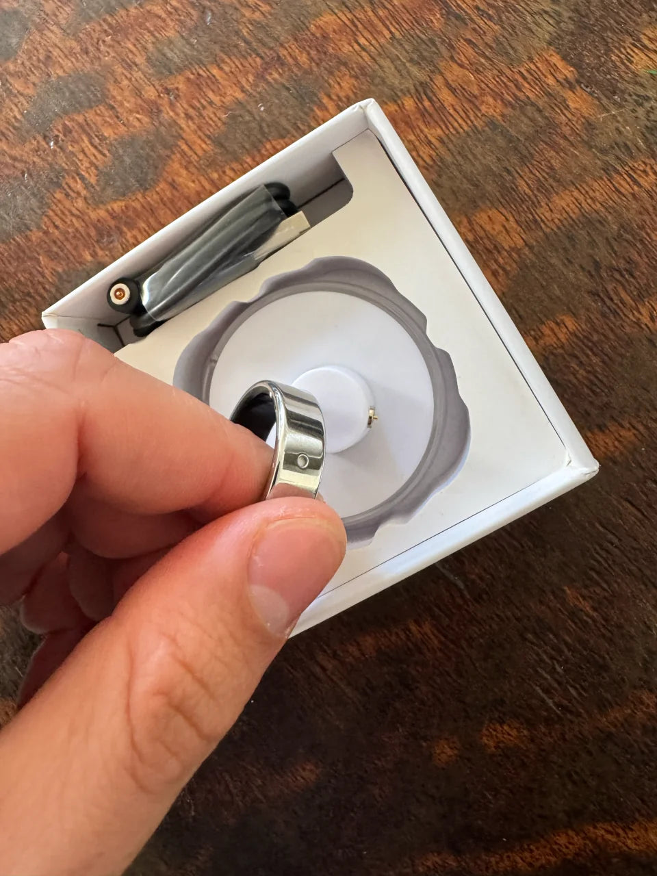 A hand holds a silver Secondly ring above its open box, showing the button.