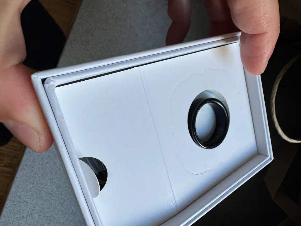 Hands hold an open white box containing a black Secondly ring.